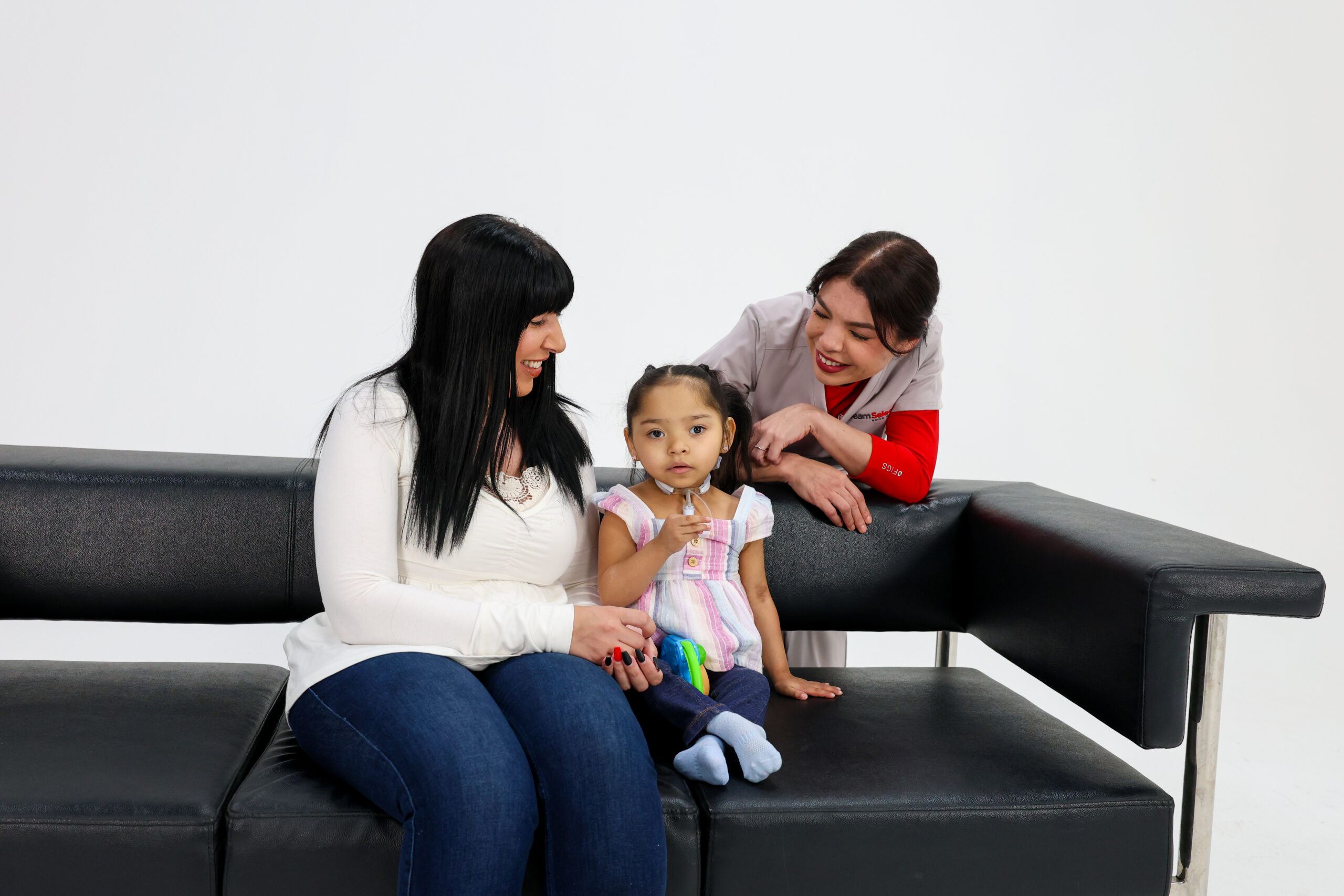 Three people sit together on a black leather couch: a mother on the left, a young girl in the center, and a caregiver in a gray uniform leaning in from the right to help.