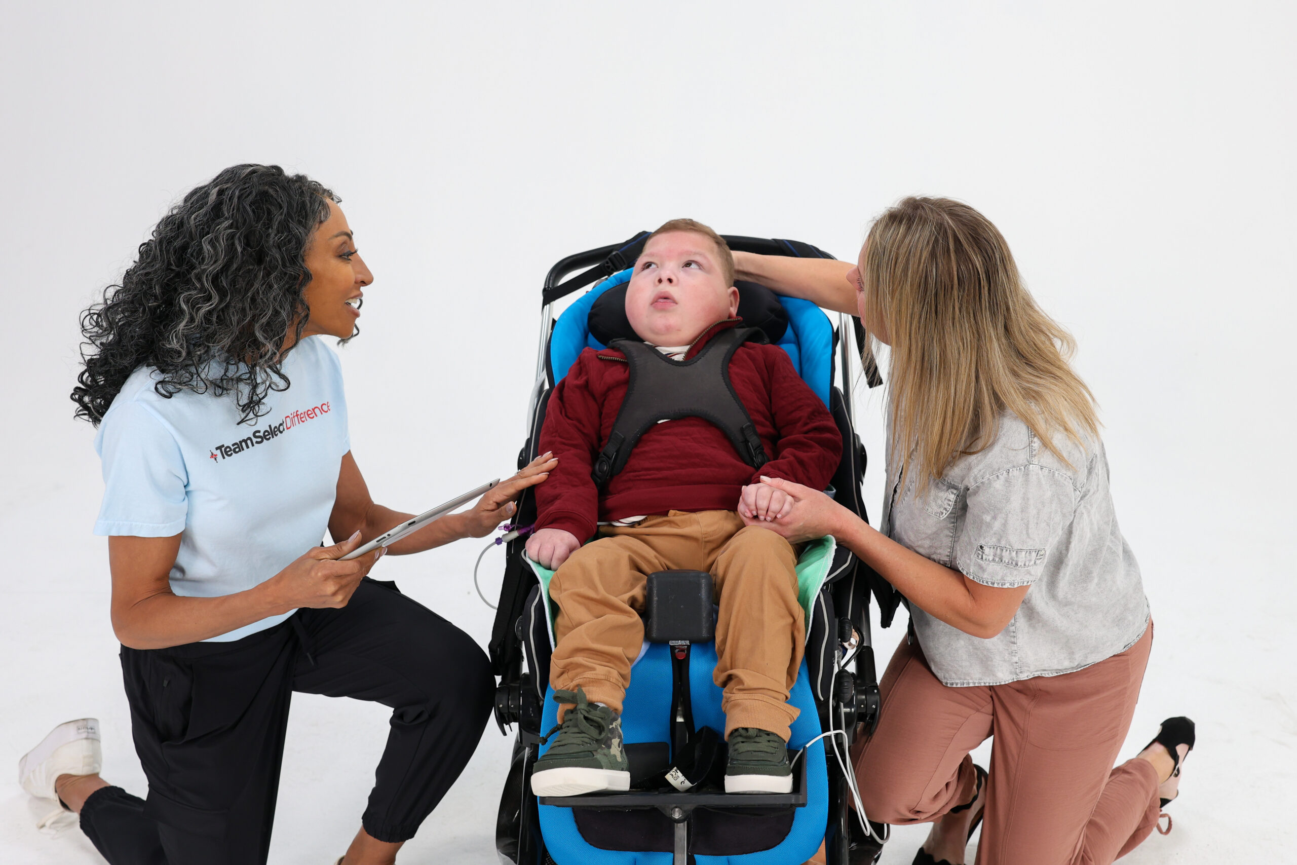 Two women kneel beside a smiling boy in a blue wheelchair, talking with him in a bright, open space.