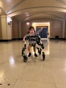 Smiling child in a gait trainer walking in a spacious indoor hallway with arched ceiling.] ,