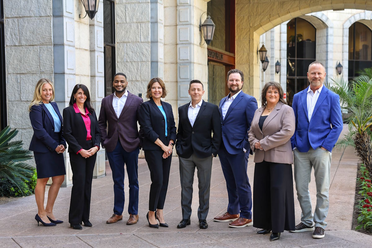 about-us Eight professionally dressed individuals pose together outdoors in a courtyard, smiling at the camera in front of a stone building.